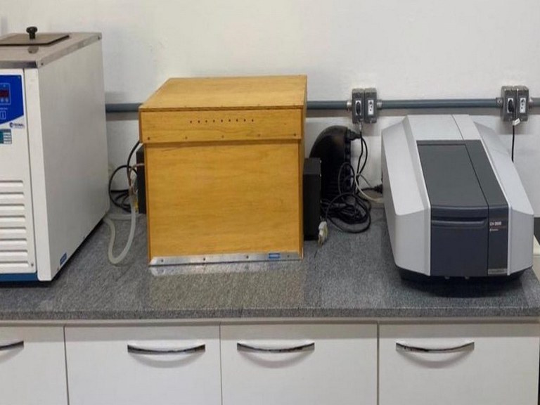 Descrição da imagem. A imagem apresenta uma fotografia de uma bancada de um laboratório com três equipamentos em cima. O primeiro da esquerda para a direita é denominado banho térmico, equipamento retangular de cor branca, com detalhes do visor em azul, apenas uma parte dele está visível na foto. No meio, uma caixa em madeira denominada de caixa de degradação. E a direita o espectrofotômetro, equipamento também retangular de cores branca e cinza, mais baixo que os outros dois. Abaixo da bancada é possível observar 5 portas de um armário branco, todas fechadas, com puxadores prateados. Ao fundo parede branca, onde tomadas e fios estão visíveis. Fim da descrição 
