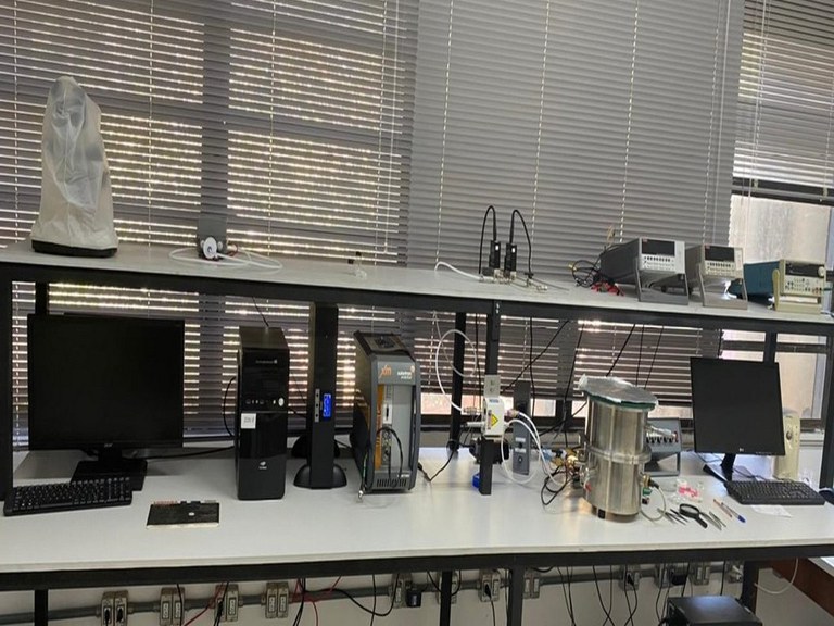 Descrição da imagem. A imagem apresenta uma fotografia de uma bancada branca com equipamentos eletrônicos. Na parte de cima é possível observar três eletrômetros, um controlador de fluxo e um microscópio coberto com uma capa plástica. Na parte de baixo é possível observar, dois monitores, dois teclados e uma Unidade Central de Processamento do computador, há também um nobreak, um impedancímetro e uma câmara para testes de detecção de gás. Ao fundo janelas com persianas cinza. Fim da descrição