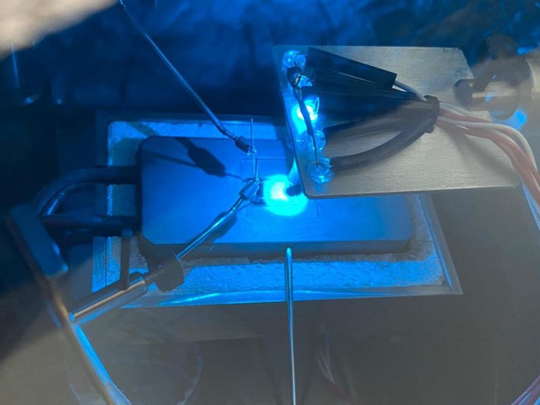 Descrição da imagem. A imagem apresenta uma fotografia de um equipamento de laboratório. No centro da imagem é possível observar um retângulo com bordas metálicas, dentro do qual há uma chapa de aquecimento também em forma retangular, sobre a qual é possível observar um ponto de luz brilhante de cor azul, o qual deixa toda a imagem em tons azulado. Acima, há um suporte para este ponto de luz, também de forma retangular e metálico, com cinco fios encapados. Fim da descrição.